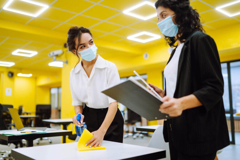 A woman is cleaning a desk in an office. She is using a cloth or sponge to wipe down the surface, removing dust and clutter. The desk is neatly organized, with office supplies and a computer arranged in an orderly fashion. The woman appears focused on her task, demonstrating a commitment to maintaining a tidy and professional workspace. The office has a bright, inviting atmosphere, emphasizing the importance of cleanliness in a work environment.