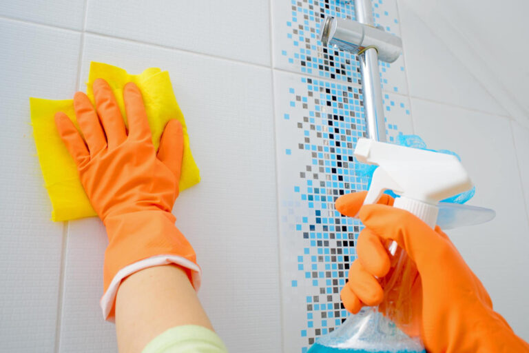 A hand wearing orange gloves is cleaning a shower. The hand is scrubbing the tiles and glass door, using a cloth or sponge to remove soap scum and water spots. The shower features modern fixtures and bright tiles, reflecting light and cleanliness. This action emphasizes the importance of maintaining a hygienic bathing area, showcasing a thorough approach to cleaning.