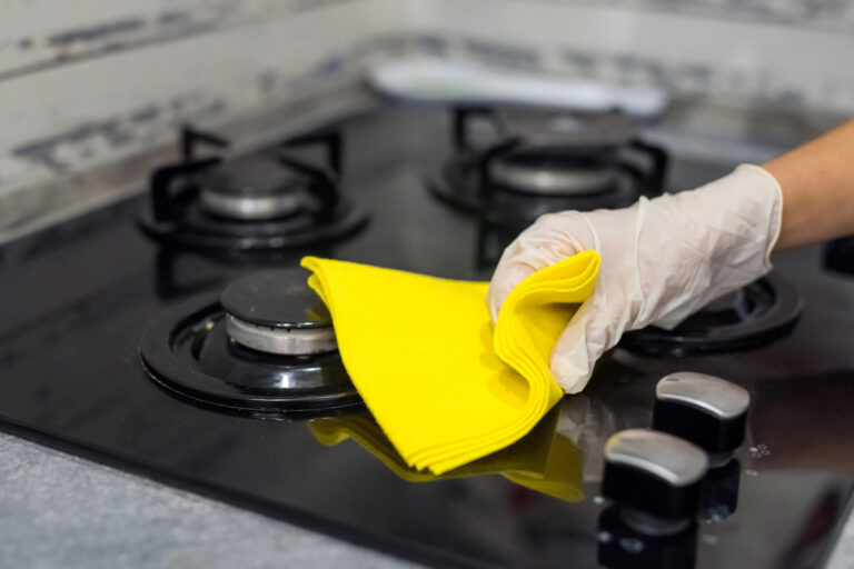 A hand wearing blue gloves is cleaning a stove. The hand moves methodically over the burners and surface, using a cloth or sponge to remove spills and grease. The stove appears sleek and modern, with shiny surfaces reflecting light. This action emphasizes the importance of cleanliness in the kitchen, showcasing a commitment to maintaining a hygienic cooking space.