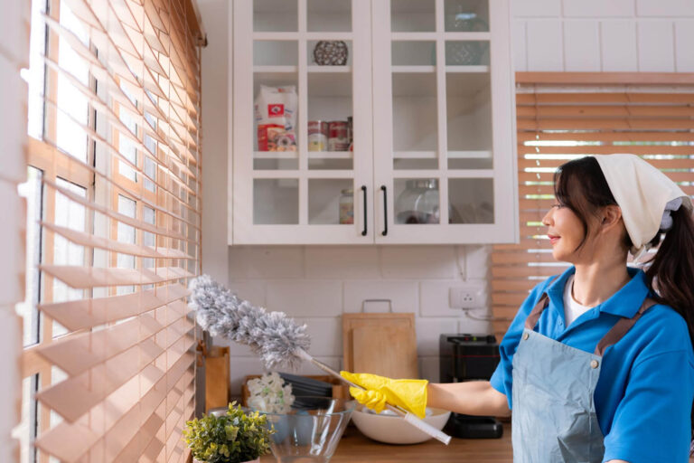 A woman is cleaning a kitchen window while wearing yellow gloves. She carefully wipes down the glass with a cloth, making sure to remove any smudges and leave the surface clear and shiny. The bright kitchen light reflects off the window as she works, emphasizing the clean, airy feel of the space. Her focus is on reaching all parts of the window, ensuring it’s spotless and enhancing the overall cleanliness of the kitchen. The best maid services in Queens