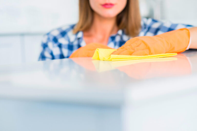 A woman is cleaning a shelf while wearing orange gloves. She carefully wipes down the shelf, ensuring that it's free of dust and dirt. The shelf is part of a neat and organized space, with items like books, decor, or storage containers nearby. Her pink gloves stand out as she moves her hand back and forth, demonstrating a focus on cleanliness and order in the area.